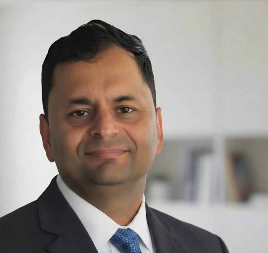 Smiling man in a dark suit and blue tie posing in a bright office setting.