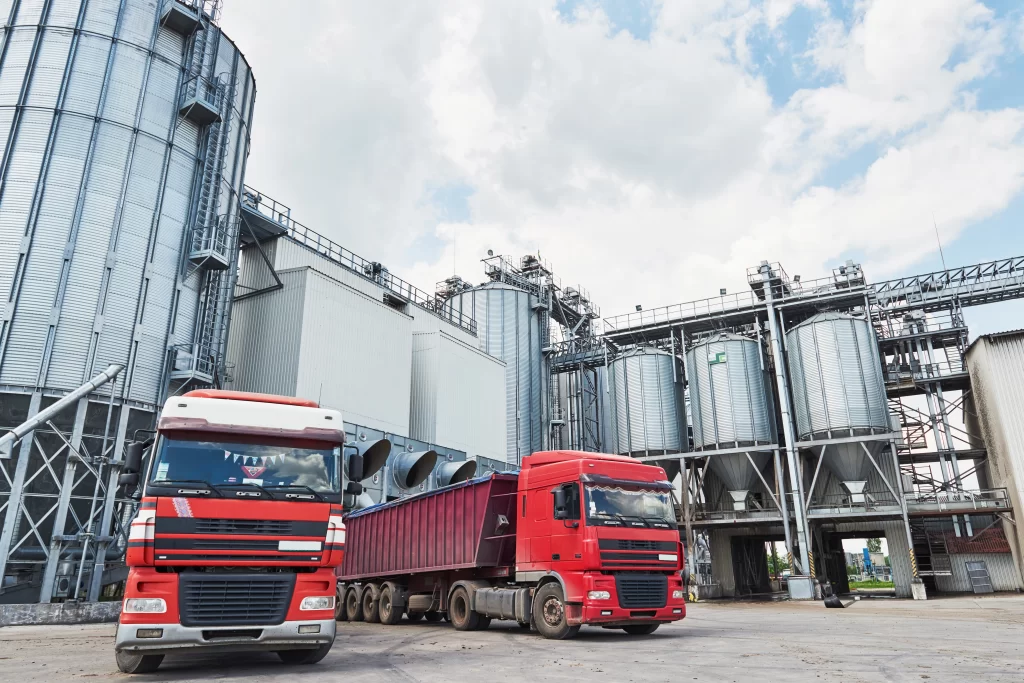 Industrial plant with tall metal silos and red cargo trucks parked in front.