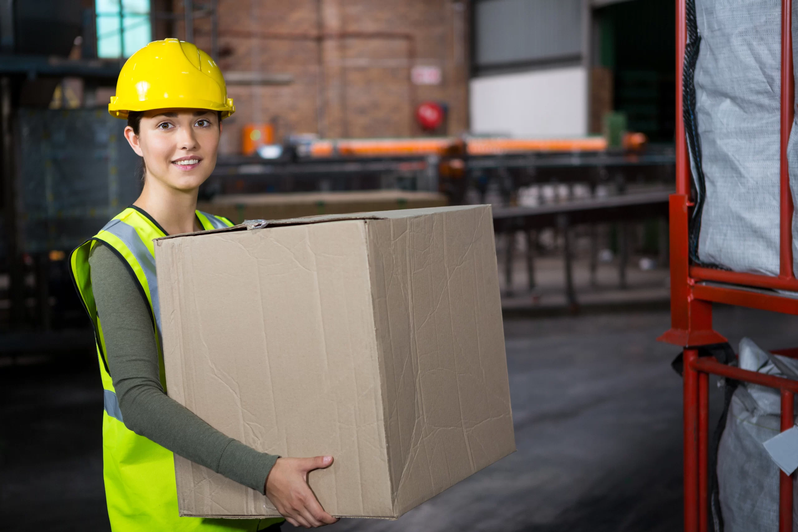 Female warehouse worker in a yellow hard hat and high-visibility vest holding a cardboard box.