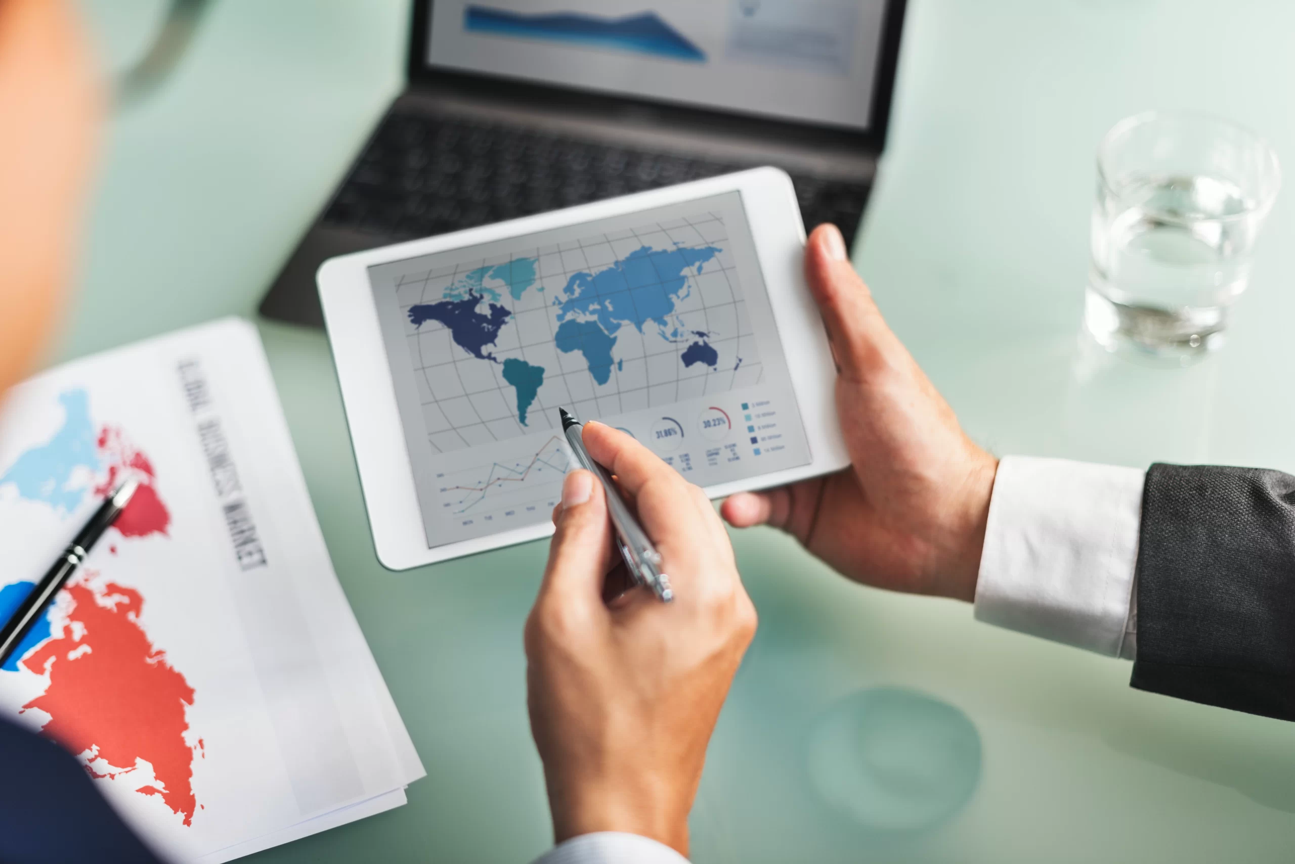 Person holding a tablet showing a world map and charts during a business meeting at a desk with papers and a glass of water