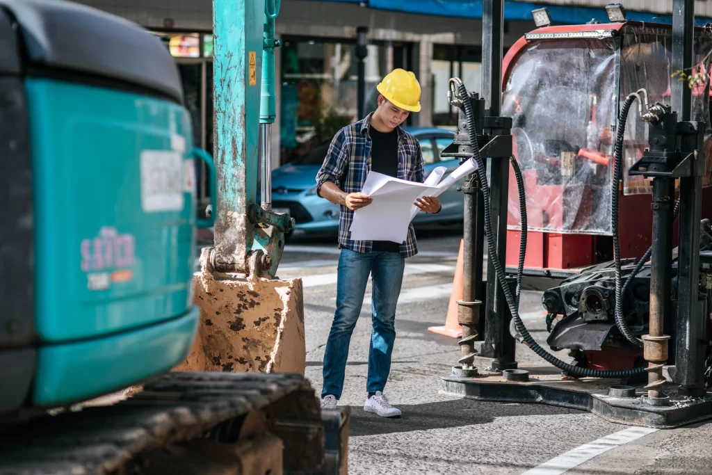 Construction worker with yellow hard hat reviewing blueprints among heavy equipment outdoors