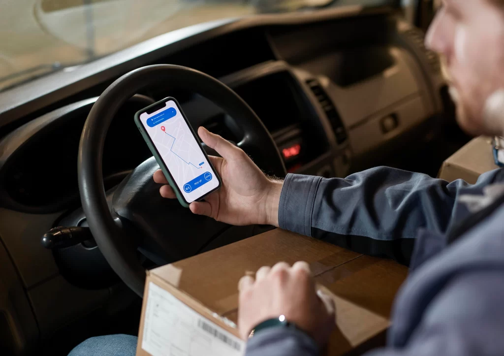 Delivery driver in a car using a smartphone map app to navigate, with a cardboard package and clipboard on the seat nearby.