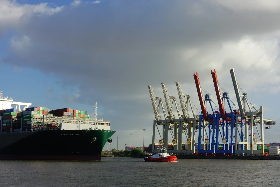 Large container ship named Ever Golden docked at a busy port with stacked colorful containers and tall port cranes in the background, a red tugboat on the water nearby.