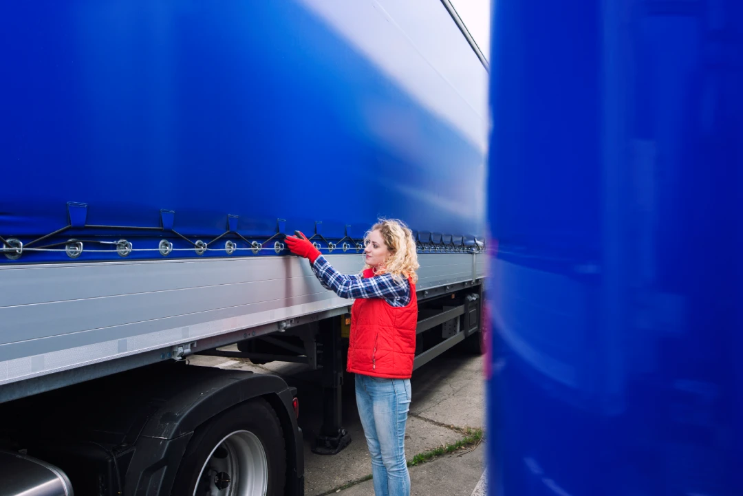 Woman in a red vest and red gloves stands beside a blue truck trailer, touching the side of the tarpaulin cover as she checks or secures it.