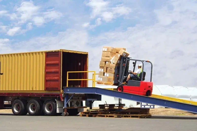Forklift moves boxes from a yellow shipping container onto a ramp at an industrial loading dock.