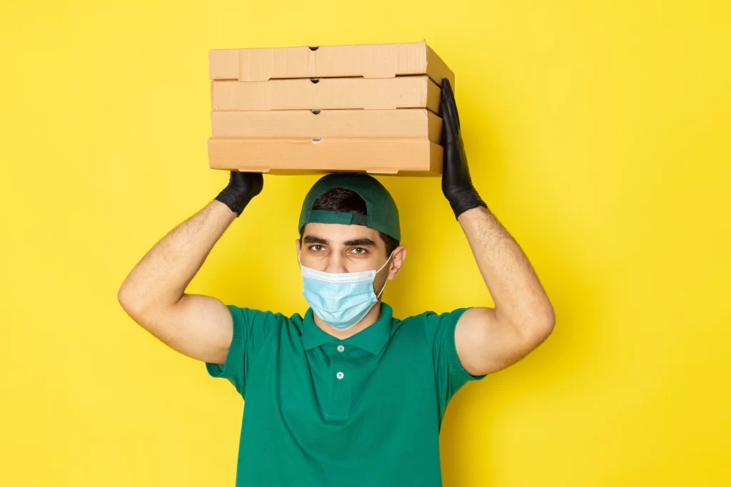 Delivery person wearing a mask and gloves balances a stack of four pizza boxes on their head against a bright yellow background.