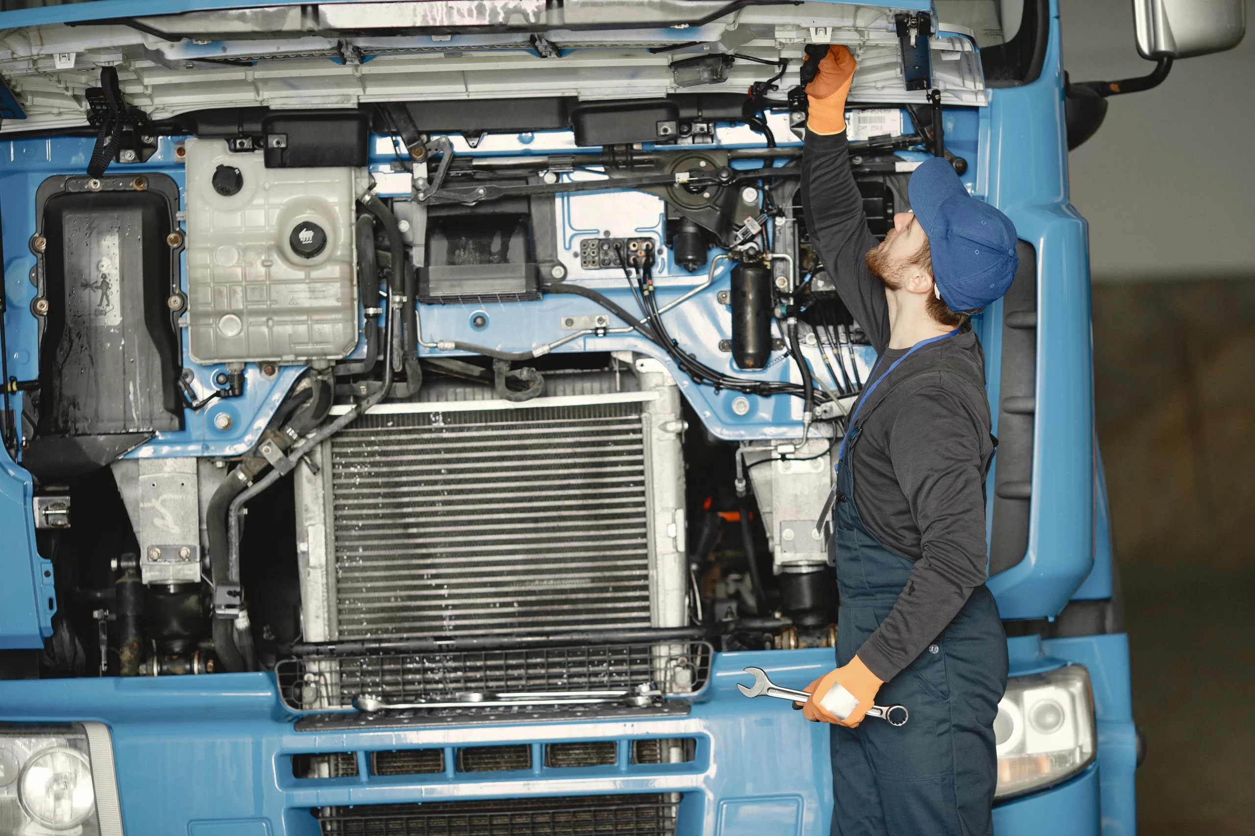 Mechanic in a blue cap and gloves works under the hood of a blue truck, inspecting the exposed engine bay.