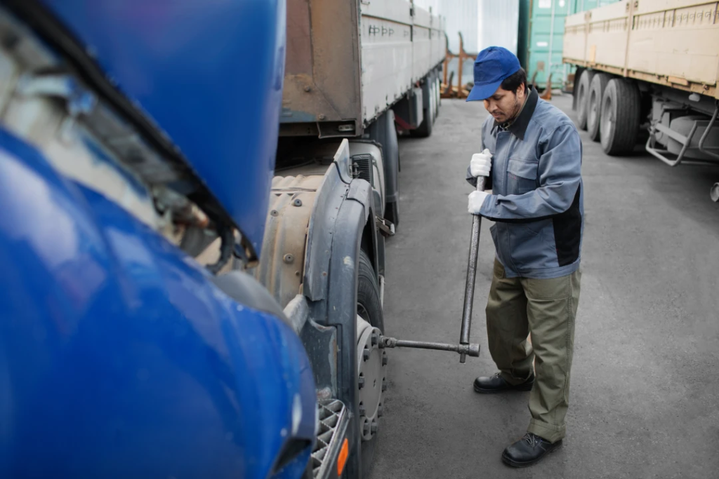 Truck maintenance worker in a blue cap tightens a wheel with a long bar beside a blue tractor-trailer in a yard by parked trucks.
