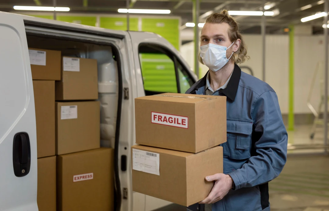 Delivery worker wearing a mask unloads fragile boxes from a white van in a loading area.