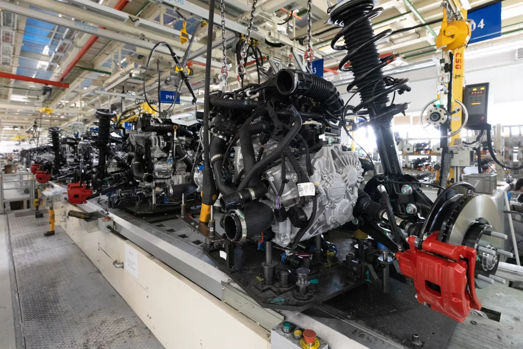 Industrial engine assembly on a production line with numerous hoses and cables overhead or suspended parts above a moving platform.