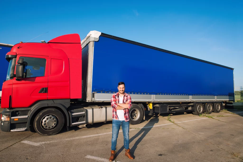 Man in plaid shirt and jeans standing with arms crossed in front of a red semi-truck with a blue trailer