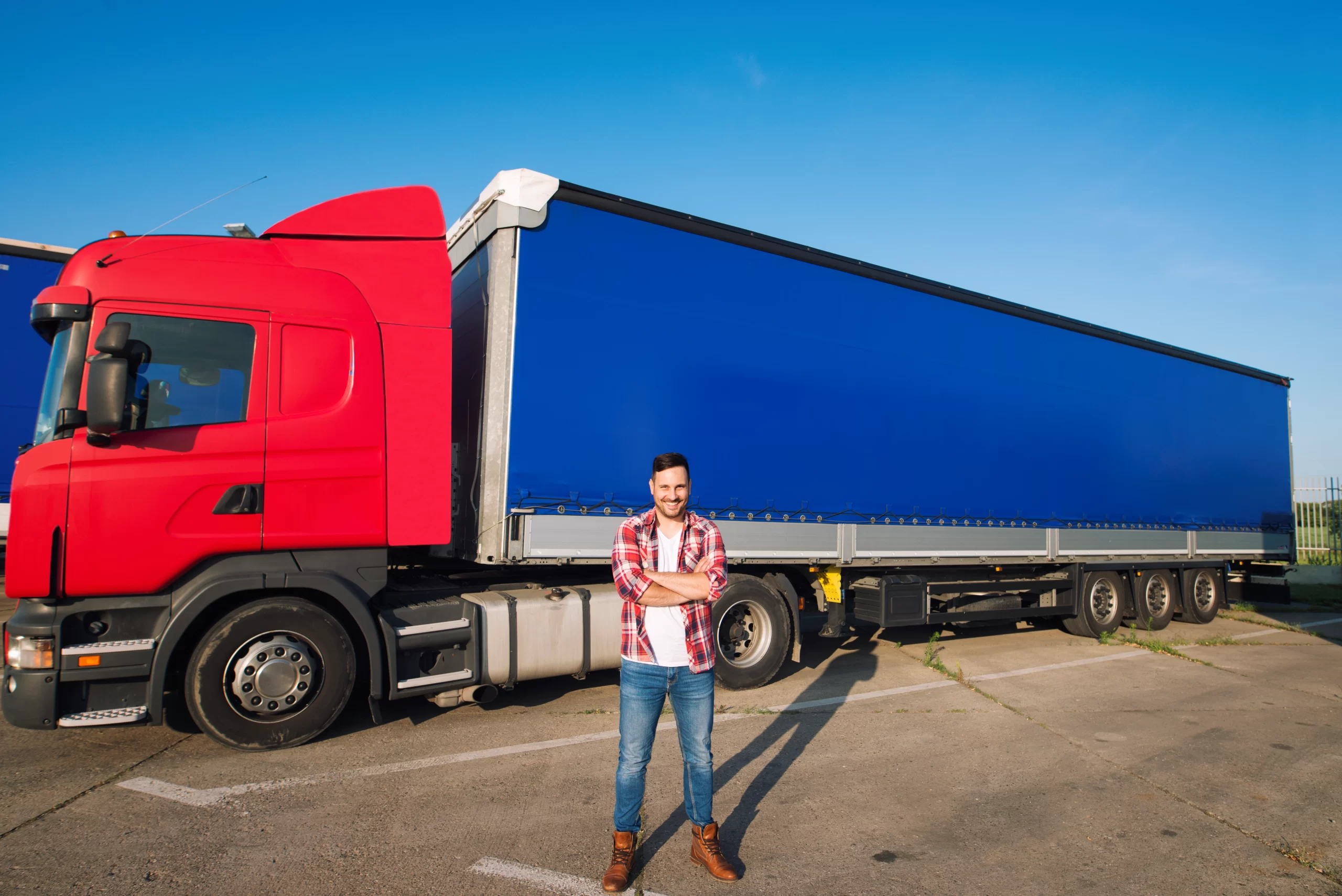 Man in plaid shirt and jeans standing with arms crossed in front of a red semi-truck with a blue trailer