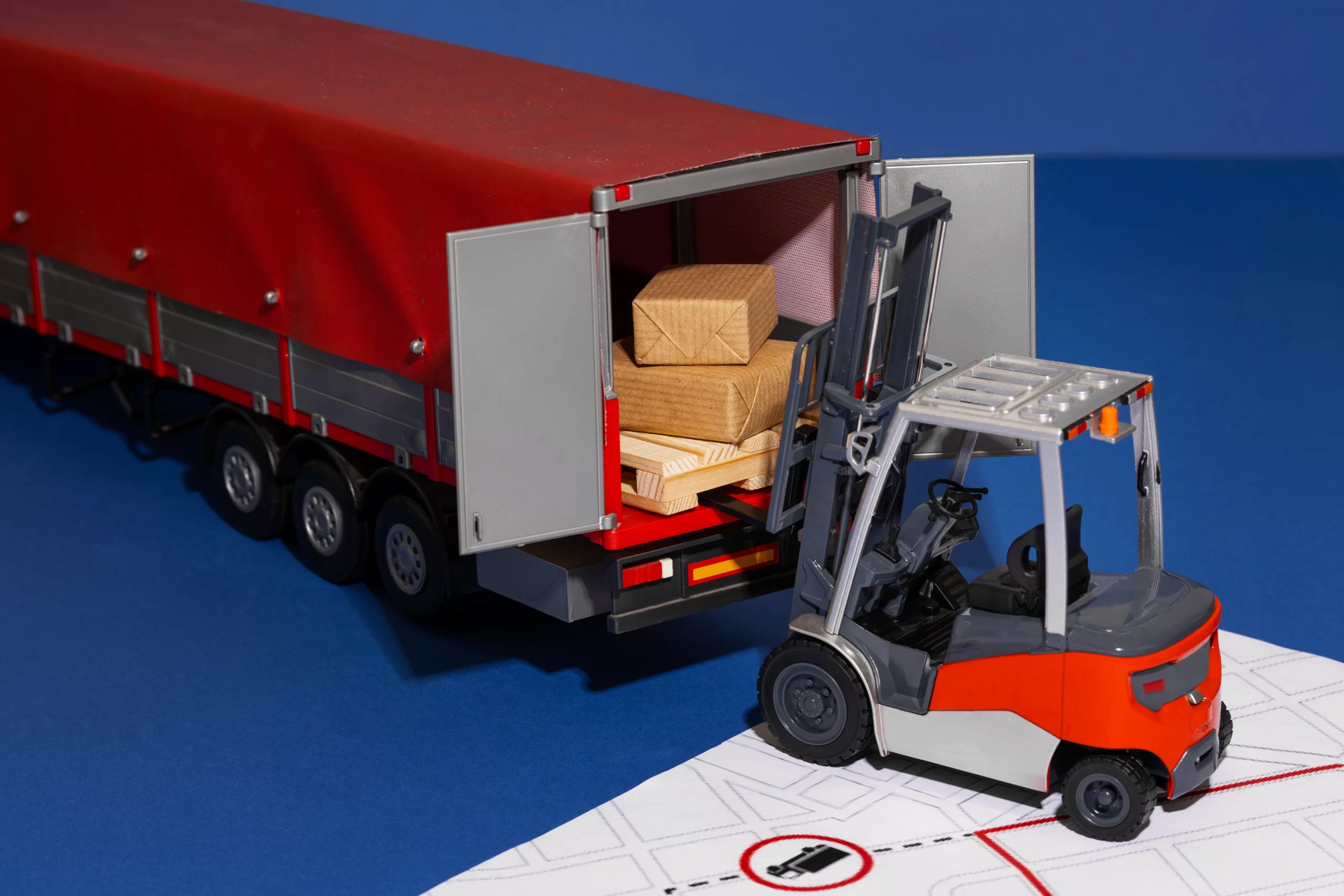 Toy forklift loading wooden boxes into a red trailer on a blue surface, showing logistics in action.