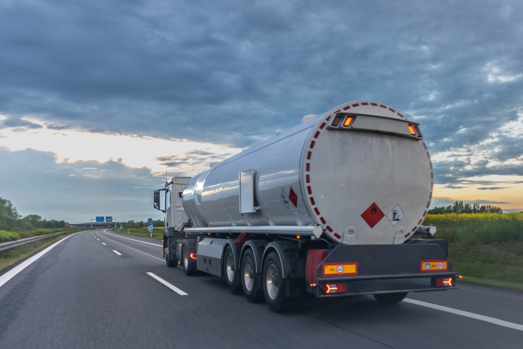 Rear and side view of a white tanker trailer driving on a multi-lane highway beneath a cloudy sky, with red hazard placards and illuminated brake lights on the back.