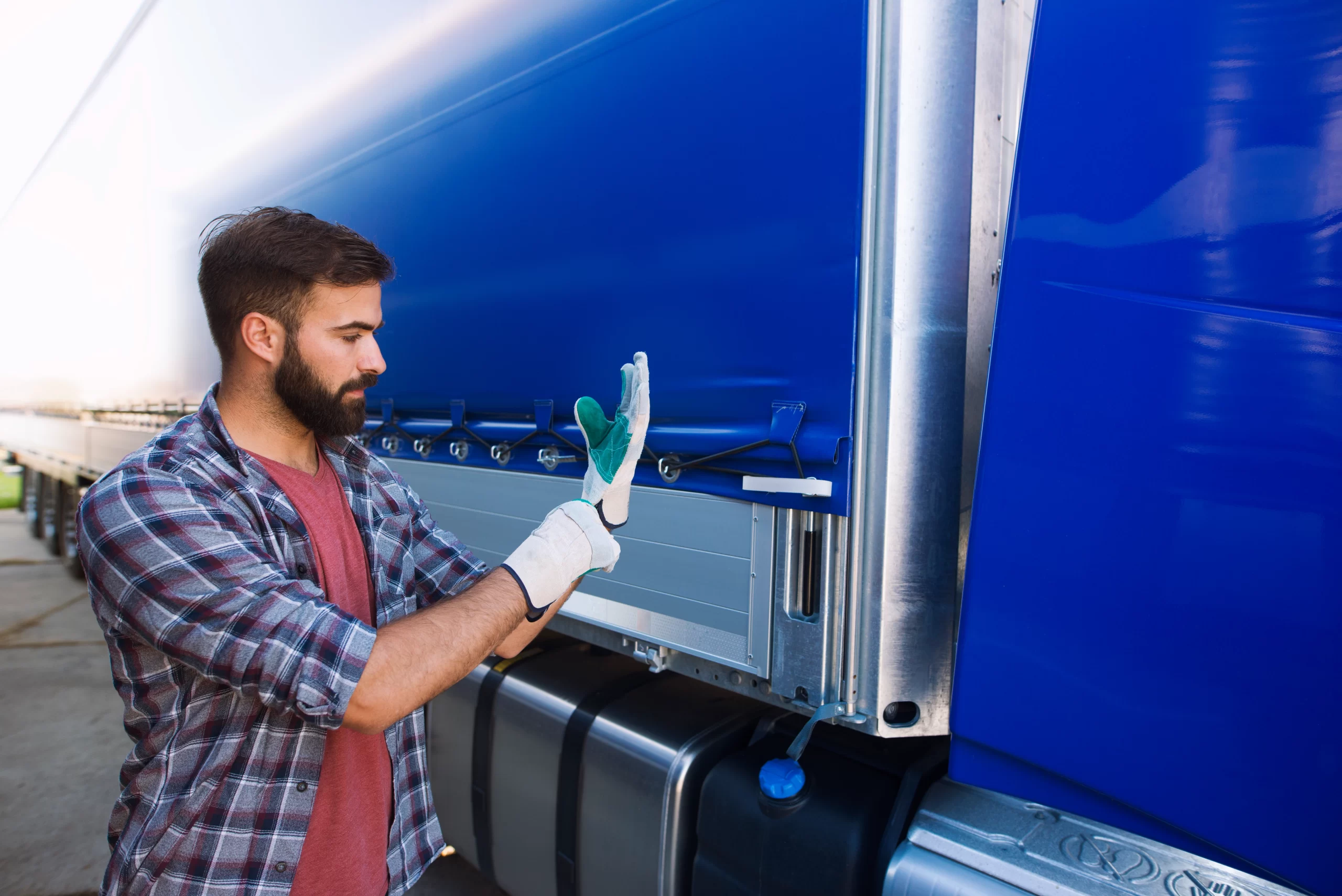 Maintenance worker in a plaid shirt and gloves inspecting the latch on a blue semi-trailer truck door.