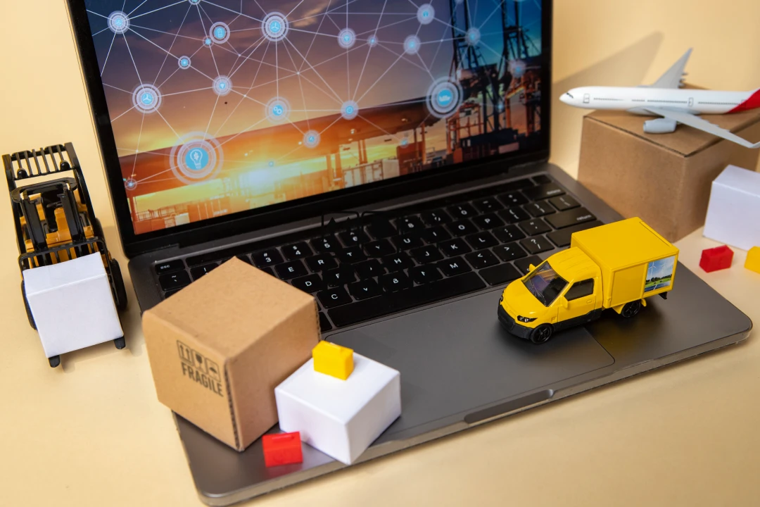 Laptop on a desk with a network-themed screen, surrounded by toy delivery trucks and shipping boxes. (logistics theme)