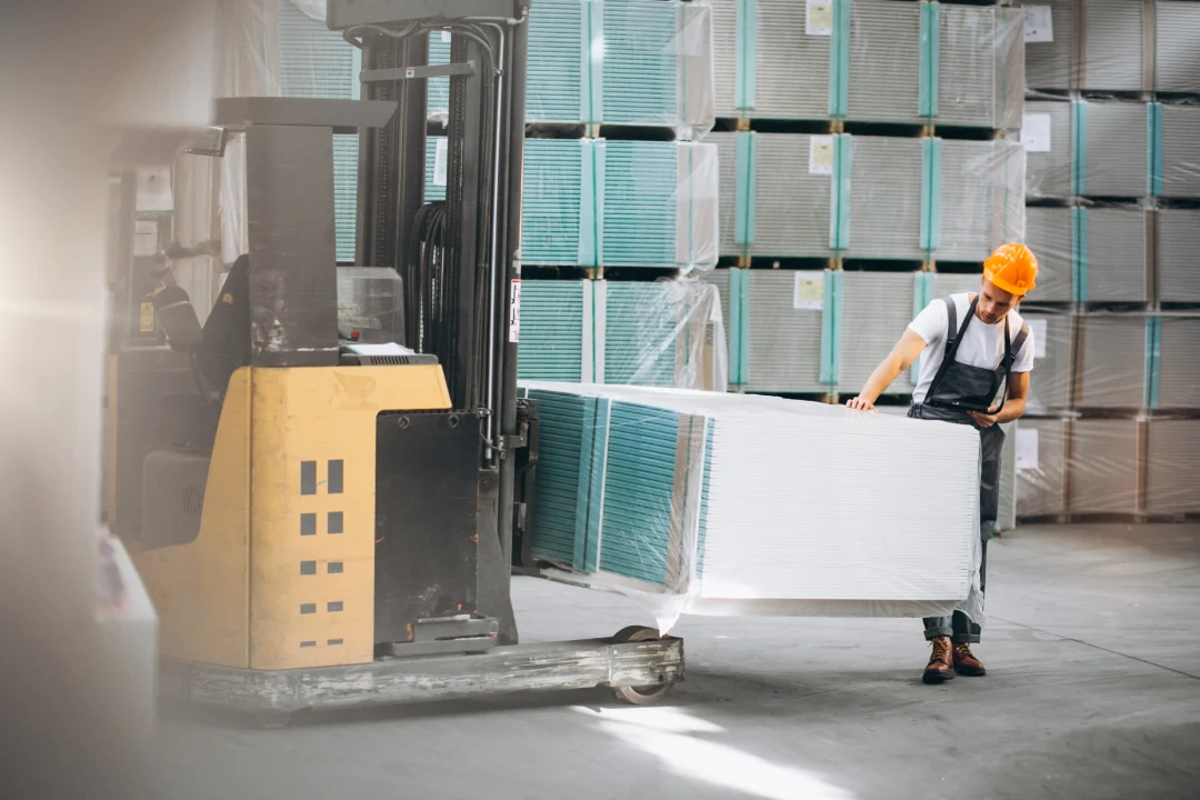 Warehouse worker in an orange hard hat guides a wrapped pallet of large white panels beside a forklift in a warehouse.