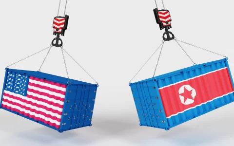 Two blue shipping containers hung by crane hooks, one painted with the American flag and the other with the North Korean flag.